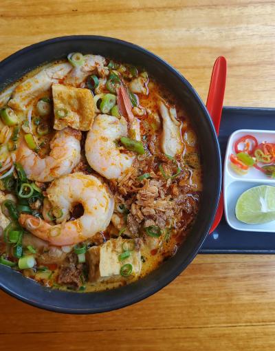 Spicy laksa broth accompanied by prawns and chicken. a small dish containing chilli and lime is to the right of the bowl.